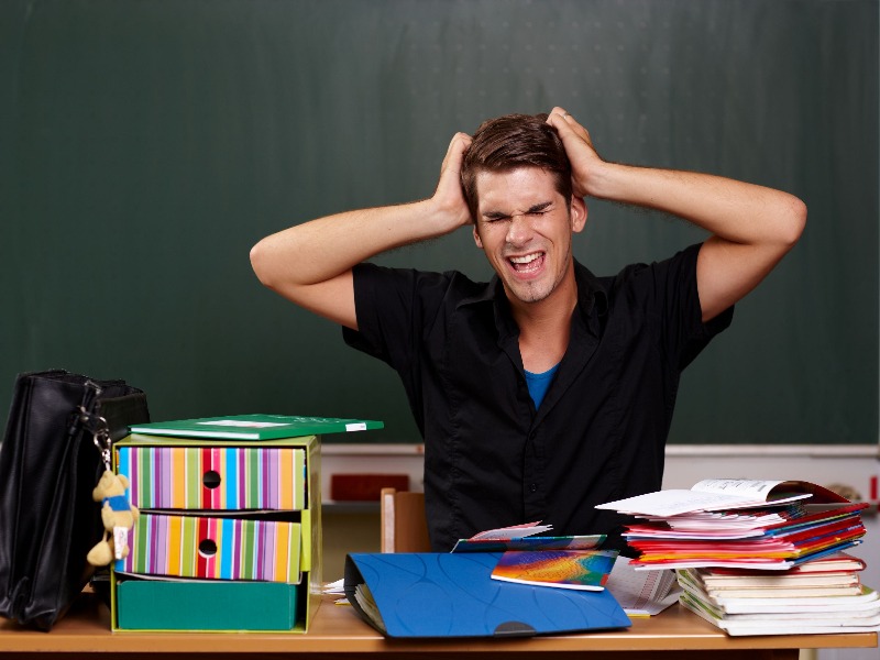 Person sitzt an einem Schreibtisch vor einer Tafel mit Händen am Kopf und mehreren Ordnern und Büchern auf dem Tisch